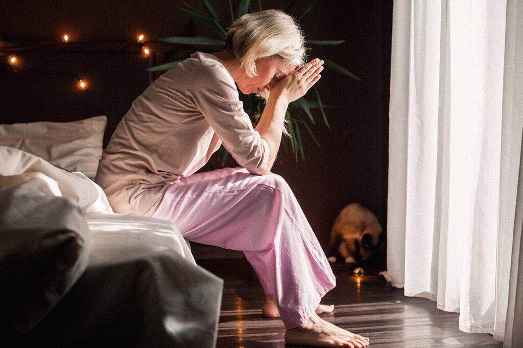 A middle-aged woman in pink pyjamas sits on the edge of her bed with her head hanging down and her hands to her forehead.