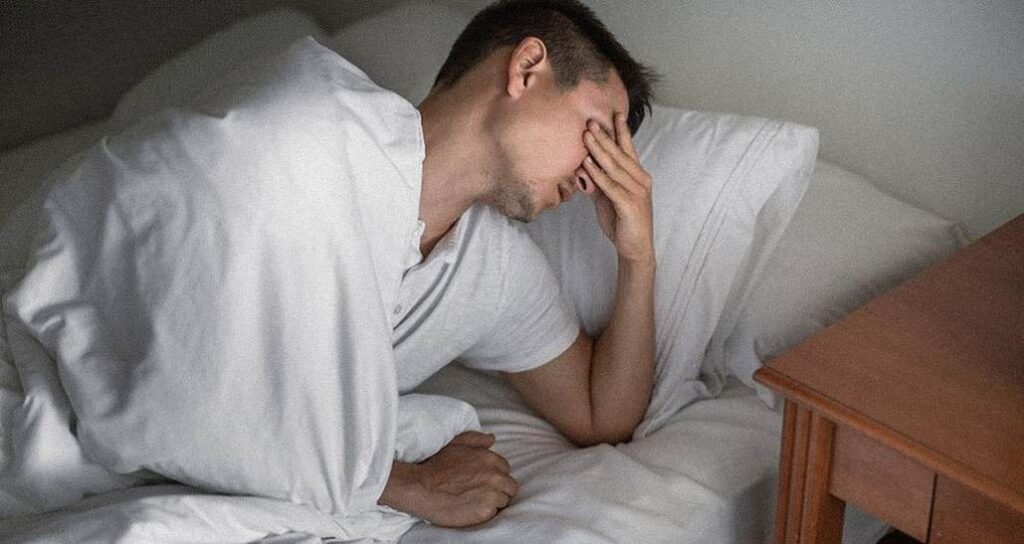 Un homme se tenant la tête après une mauvaise nuit de sommeil et ayant du mal à sortir du lit.