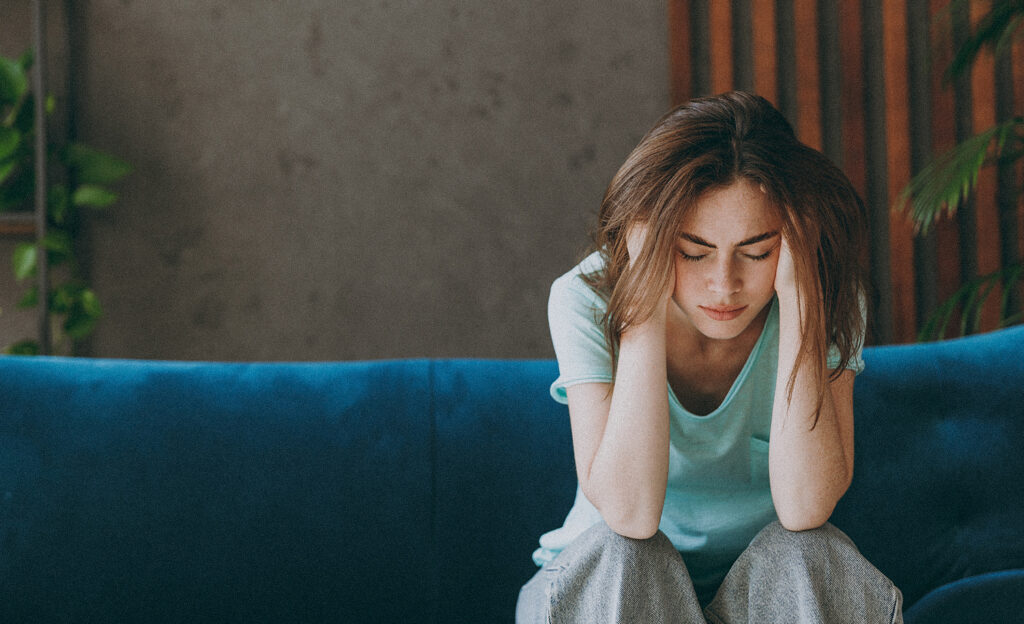 Femme dans la trentaine, épuisée, vêtue de vêtements décontractés, se tenant la tête dans les mains, les yeux fermés en signe de fatigue.