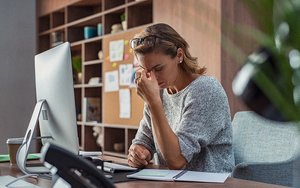 A business woman at her desk with a computer in front of her in an office. She is holding her head to signify a headache, tiredness, and stress.