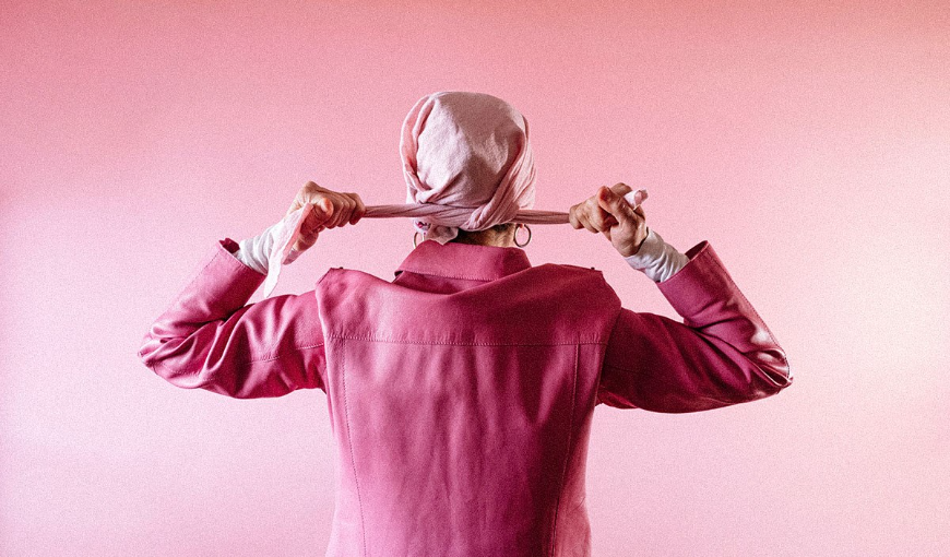 A woman against a pink backdrop tying a pink scarf around her head to fight breast cancer.